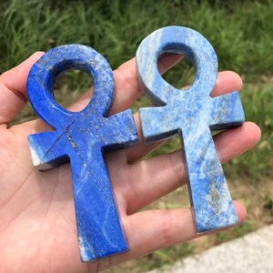 May include: Two blue lapis lazuli ankh pendants. The ankhs are carved from stone, with a cross shape and a loop at the top. The stone has a mix of dark and light blue hues with flecks of gold.