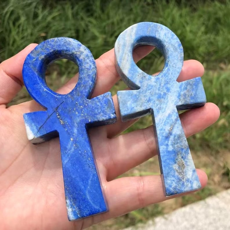 May include: Two blue lapis lazuli ankh pendants. The ankhs are carved from stone, with a cross shape and a loop at the top. The stone has a mix of dark and light blue hues with flecks of gold.