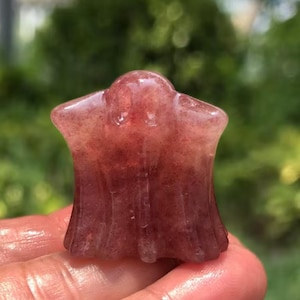 May include: A translucent, reddish-pink crystal carving in the shape of a ghost. The crystal has a smooth, polished surface and a slightly textured appearance. The ghost figure has a rounded head and a flowing, draped body.