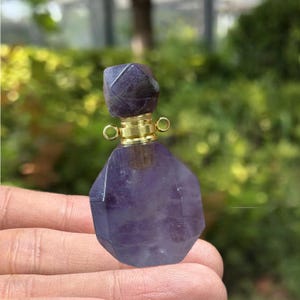 Puede incluir: Un frasco de perfume de amatista púrpura facetada con un cuello y tapón de metal dorado. El frasco tiene una forma geométrica y está sostenido en una mano. El fondo es follaje verde borroso.