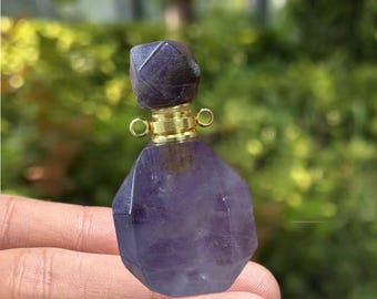 May include: A faceted, purple amethyst perfume bottle with a gold-colored metal collar and stopper. The bottle has a geometric shape and is held in a person's hand. The background is blurred green foliage.