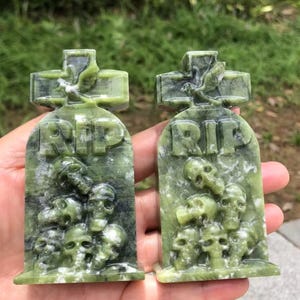 May include: Two green stone tombstone sculptures with the letters "RIP" carved into them. The tombstones have a cross at the top and are decorated with skulls. The sculptures are made of a green and white marbled stone.