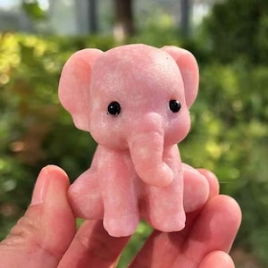 May include: A small, pink elephant figurine with black eyes and a trunk. The elephant is carved from a smooth, possibly stone-like material. The figurine is held in a person's hand, with a blurred green background.