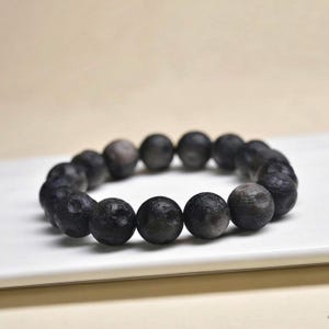 Könnte beinhalten: Ein kreisförmiges Armband, das aus zahlreichen kugelförmigen Perlen mit einer dunklen, matten und strukturierten Oberfläche besteht. Die Perlen sind einheitlich groß und in einer durchgehenden Schleife angeordnet, in Anthrazit bis Schwarz.
