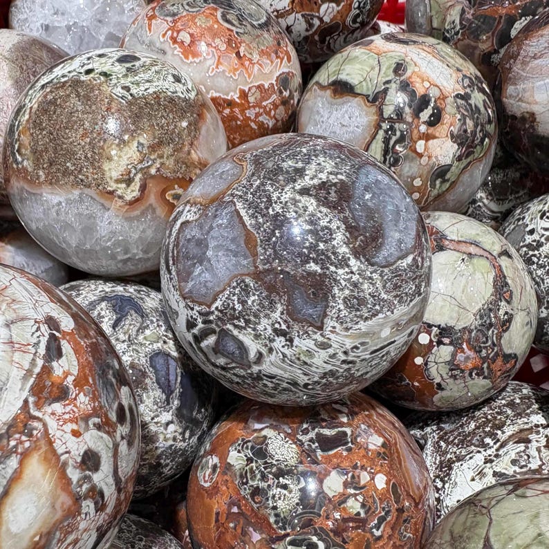 May include: A collection of polished stone spheres in various sizes and colors. The spheres display a mix of brown, gray, white, and green patterns, with some having a marbled appearance. The overall aesthetic is natural and earthy.