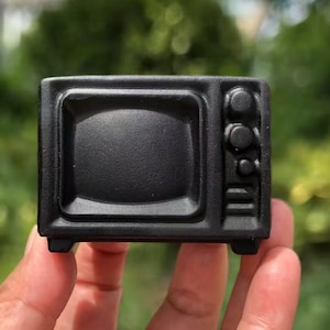 May include: A small, black, miniature microwave oven. The microwave has a rectangular shape with a rounded rectangular screen, three control knobs, and a small base with four feet. The object is held in a person's hand.
