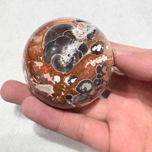 May include: A polished orb of jasper stone, featuring a mix of reddish-brown, black, and white patterns. The sphere is held in a hand, showcasing its smooth, rounded form. This decorative item is ideal for home decor or as a unique gift.