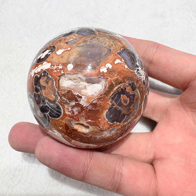 May include: A polished sphere with a unique pattern of brown, white, and black markings. The orb has a smooth, glossy surface and a mix of earthy tones. The sphere is held in a hand.