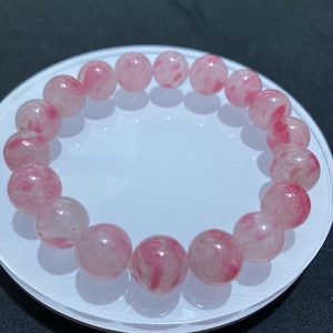 Puede incluir: Un brazalete con esferas de cuarzo rosa translúcido con vetas rojas. El brazalete está ensartado en una banda elástica, y las cuentas tienen aproximadamente 2 cm de diámetro. El brazalete se exhibe sobre una superficie blanca.