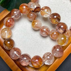 Puede incluir: Un brazalete de cuentas esféricas translúcidas con inclusiones internas. Las cuentas muestran una mezcla de colores cálidos, incluyendo naranja, marrón y tonos claros. El brazalete se presenta sobre una bandeja de madera, mostrando la belleza natural de las piedras.