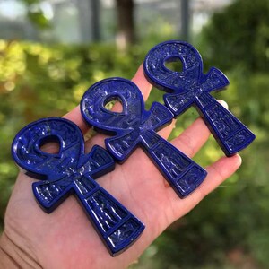 May include: Three lapis lazuli ankh pendants are displayed. The pendants are a deep blue color with carved details. The ankhs are held in a hand against a blurred green background.