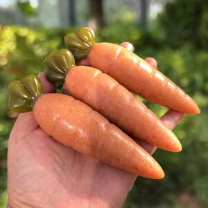 May include: Hand holding three orange carrot-shaped sculptures with green tops. The carrots have a smooth, polished appearance, and the green tops have a textured, leafy design. The background is blurred, suggesting an outdoor setting.