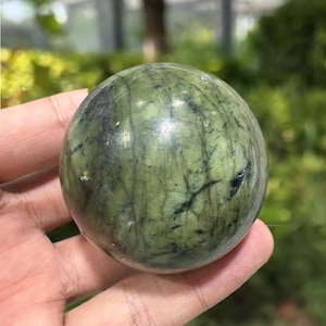 May include: A green and black stone sphere. The sphere has a smooth, polished surface with natural veining patterns in shades of green and black. The sphere is held in a hand, with a blurred green background.