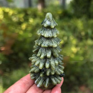 May include: A carved stone figurine in the shape of a coniferous tree. The object features a layered, pointed design with a smooth, polished surface in shades of mottled olive green, dark forest green, and subtle greyish-white veining throughout.