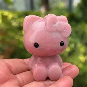 Puede incluir: Una figura rosa de gato con un lazo, ojos negros y un cuerpo redondeado. La figura se sostiene en una mano, con un fondo verde borroso.