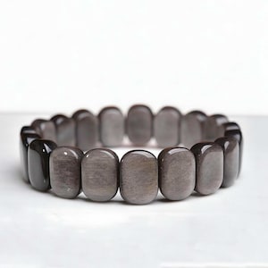 Può includere: Un braccialetto elastico composto da perline lisce, levigate e rettangolari con bordi arrotondati. Le perline presentano una tavolozza di colori grigio e antracite cangianti con sottili variazioni di tono e riflessi di luce.