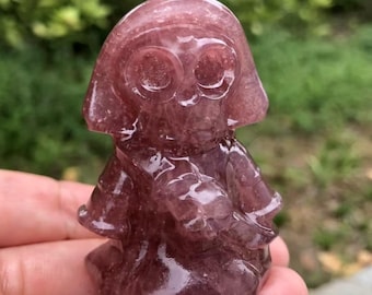 May include: A carved, reddish-purple crystal figurine of a stylized figure with large, round eyes and a hooded head. The figure is holding something in its hands, with a smooth, polished surface. The background is blurred green foliage.