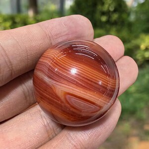 Pode incluir: Uma pedra preciosa polida e redonda com um padrão de redemoinho de faixas castanho-avermelhadas e brancas. A pedra é segurada em uma mão, mostrando sua superfície lisa e reflexiva. A pedra natural tem um acabamento brilhante.
