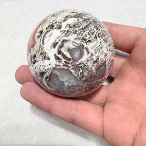 May include: A polished, spherical stone with a unique pattern of white, brown, gray, and reddish-orange hues. The orb is held in a hand, showcasing its natural textures and colors. The stone's surface has a marbled appearance.