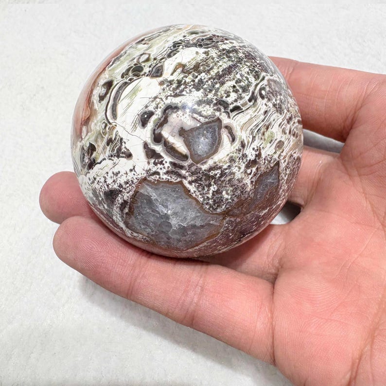 May include: A polished, spherical stone with a unique pattern of white, brown, gray, and reddish-orange hues. The orb is held in a hand, showcasing its natural textures and colors. The stone's surface has a marbled appearance.