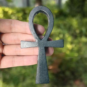 May include: A dark gray, metallic ankh symbol featuring a smooth, polished surface with a subtle speckled texture. The object has a classic cross shape with a rounded loop at the top, a horizontal crossbar, and a vertical base that tapers slightly.