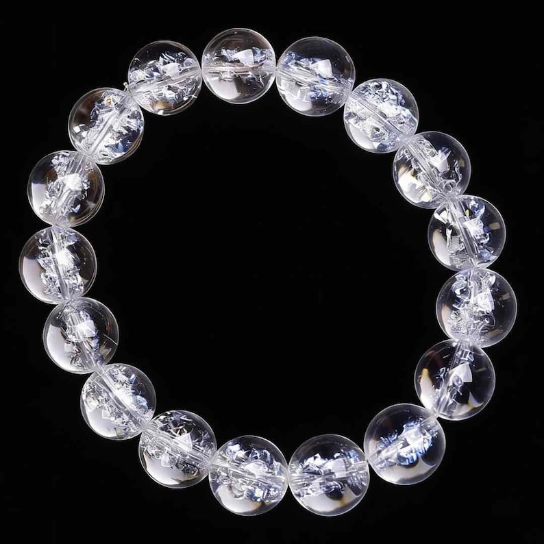 May include: A clear quartz crystal bead bracelet. The bracelet features large, round, transparent beads with internal fractures, strung together on a clear elastic cord. The beads are approximately 1 inch in diameter. The background is solid black.