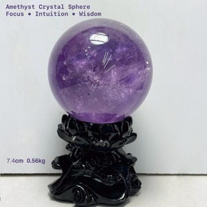 May include: A purple amethyst crystal sphere, 7.4cm in diameter, presented on a black decorative stand. The sphere has a translucent appearance with internal patterns. Text at the top reads "Amethyst Crystal Sphere, Focus, Intuition, Wisdom". The sphere weighs 0.56kg.