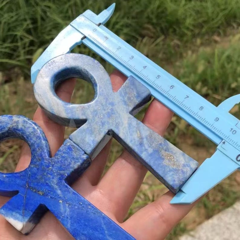 May include: Two blue lapis lazuli ankh pendants with a light blue ruler measuring one of the ankhs. The ankhs are a deep blue color with lighter blue and gold flecks. The ruler is measuring the ankh's length.