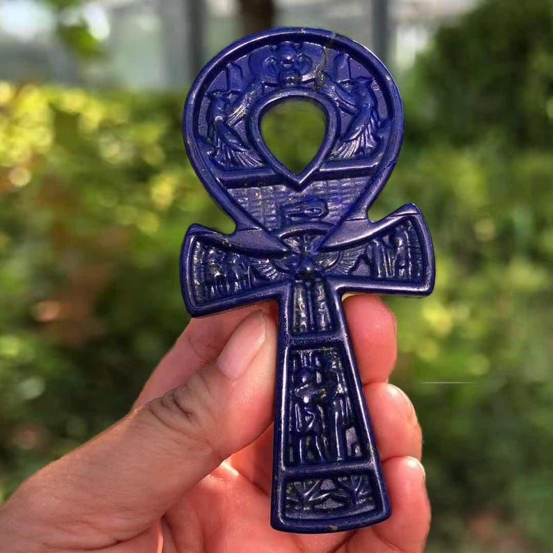 May include: A deep blue ankh pendant carved with intricate Egyptian hieroglyphs and bird motifs. The ankh, a symbol of life, features a loop at the top and a cross-like shape below. The pendant is held in a hand against a blurred green background.