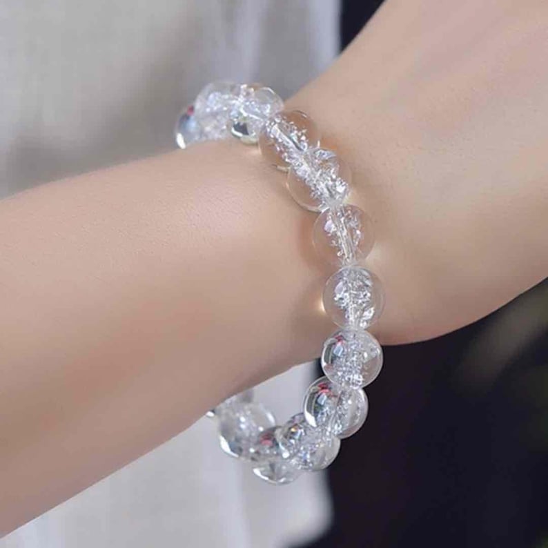 May include: A stretch bracelet composed of round, clear, crackled glass beads. Each bead features an internal fractured texture that catches the light, creating a shimmering, translucent appearance throughout the circular accessory.