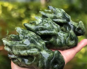May include: Three carved jade dragon heads in varying sizes, each with a detailed, textured surface and a glossy finish. The dragons are a deep green color, with darker green veining throughout. The heads are held in a hand against a blurred green background.