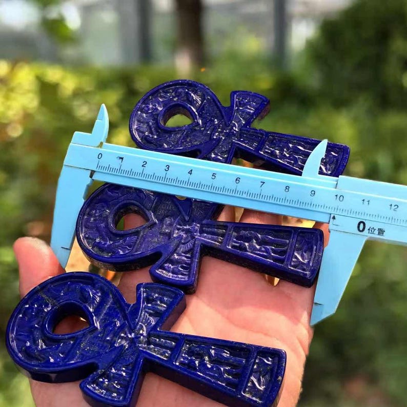 May include: Three deep blue ankh pendants, each with intricate carved details, are held near a blue ruler. The ankhs are approximately 4 inches long. The background is blurred, suggesting an outdoor setting.