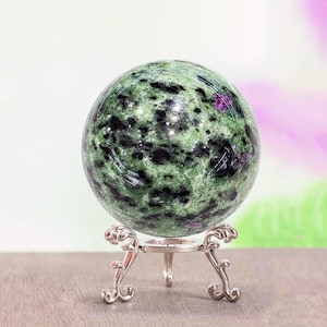 Op de afbeelding: Een groen-zwarte bol, mogelijk een edelsteen, staat op een decoratieve zilveren standaard. De bol heeft een gespikkeld patroon met hints van paars. De standaard heeft een sierlijk, opgerold ontwerp.