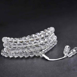 May include: A clear quartz crystal beaded bracelet, coiled and resting on a dark surface. The beads are faceted, reflecting light, and strung together with a white cord. The bracelet has a tassel of crystal beads.