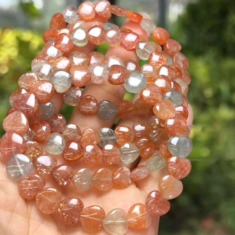 May include: A collection of heart-shaped gemstone beads in shades of orange, red, and clear. The beads are strung together to form a bracelet or necklace. The gemstones have a translucent quality, with visible inclusions. The jewelry is held in a hand.