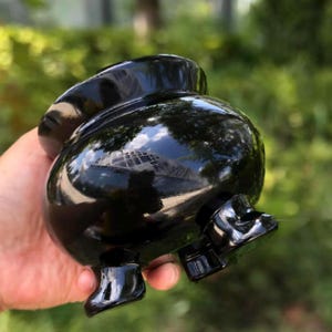May include: A polished black stone cauldron with a rounded body and a wide opening. The surface is highly reflective, showing light and environmental reflections. It features three short, sturdy legs at the base, providing a stable foundation.