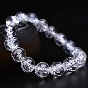 May include: A bracelet composed of round, clear, translucent beads with internal fractures and inclusions. The beads are strung together in a circular arrangement, reflecting light across their polished, spherical surfaces.