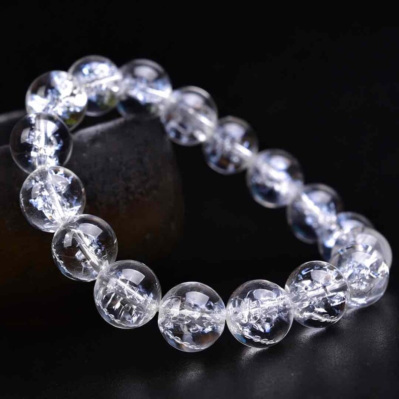 May include: A bracelet composed of round, clear, translucent beads with internal fractures and inclusions. The beads are strung together in a circular arrangement, reflecting light across their polished, spherical surfaces.