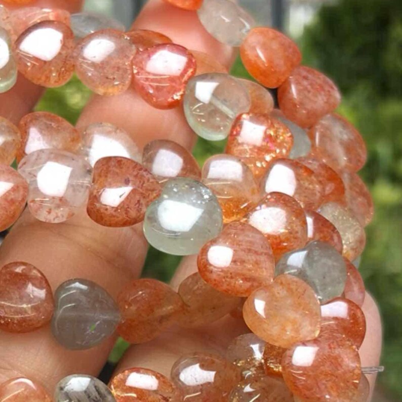 May include: Close-up of a bracelet featuring heart-shaped beads in shades of orange, red, and clear. The beads have a translucent quality, with visible internal patterns. The bracelet is held in a hand, with a blurred green background.