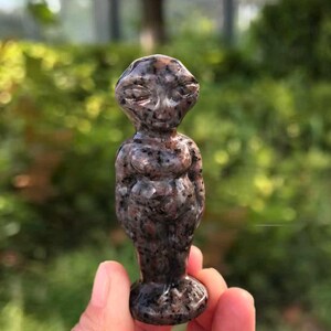 May include: A small, carved stone figurine of an alien-like figure. The statue is a mix of brown and black with a speckled pattern. The figure has a rounded head, closed eyes, and a stylized body. The base is circular.