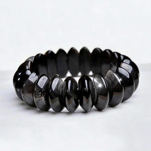 Op de afbeelding: Een elastische armband bestaande uit talloze gepolijste, zwarte, schijfvormige kralen. De kralen hebben een glad, reflecterend oppervlak met subtiele grijze insluitingen in het donkere steenmateriaal, gerangschikt in een cirkelvorm.