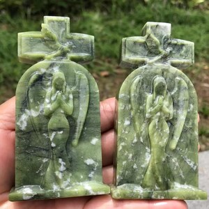 May include: Two green stone angel sculptures. Each sculpture features an angel with folded hands, wings, and a cross at the top. The stone has white veining throughout.