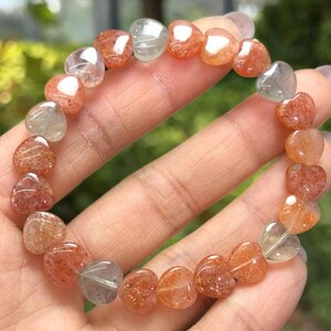 May include: A heart-shaped gemstone bracelet. The bracelet features a variety of translucent, heart-shaped beads in shades of orange, peach, and clear. The beads are strung together to form a complete circle. The bracelet is displayed on a hand.