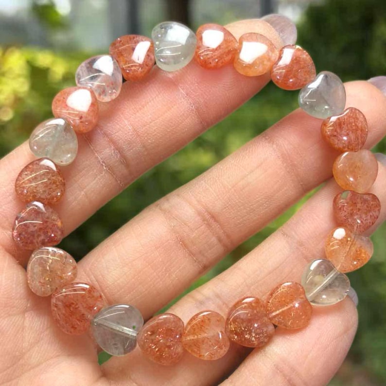 May include: A heart-shaped gemstone bracelet. The bracelet features a variety of translucent, heart-shaped beads in shades of orange, peach, and clear. The beads are strung together to form a complete circle. The bracelet is displayed on a hand.