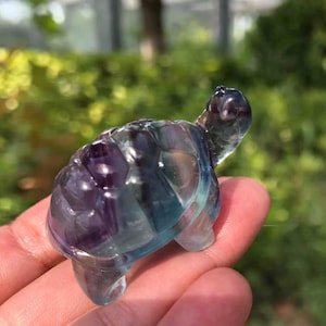 Puede incluir: Una figura de tortuga de cristal tallada. La tortuga es una mezcla de colores morado, azul y transparente. La figura se sostiene en una mano, con un fondo verde borroso.