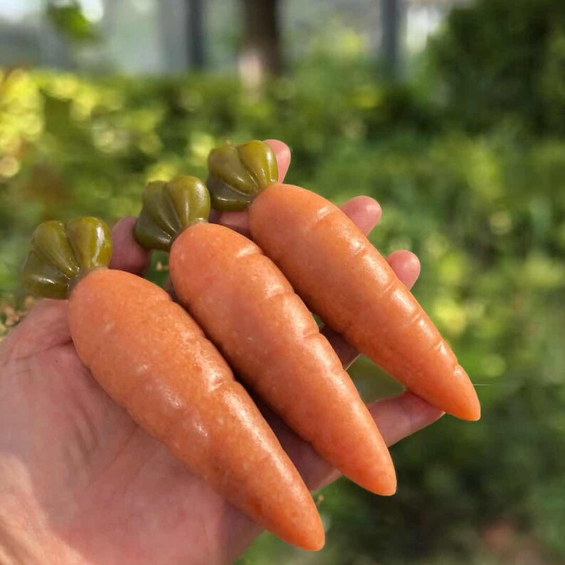May include: Three orange carrot-shaped objects with green tops are held in a hand. The carrots have a smooth, glossy appearance. The background is blurred, with green foliage.