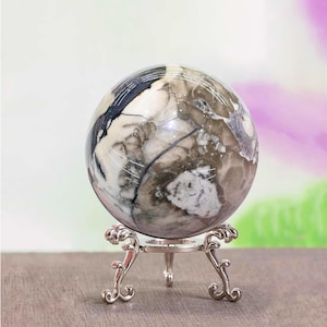 Puede incluir: Una esfera decorativa con un patrón veteado en tonos blancos, grises y negros, apoyada sobre un soporte de metal plateado. El soporte tiene un diseño ornamental y enrollado. La esfera es un punto focal, lo que sugiere un artículo decorativo o de colección.