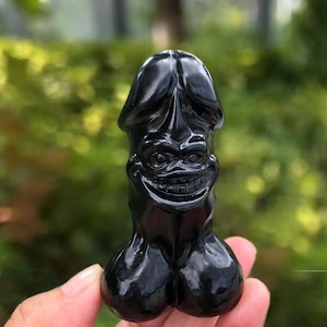 Könnte beinhalten: Eine schwarze, geschnitzte Figur mit einem lächelnden Gesicht und phallischer Form. Die Skulptur hat eine glänzende Oberfläche und wird vor einem verschwommenen grünen Hintergrund gehalten. Das Gesicht hat detaillierte Merkmale, einschließlich Augen und einem breiten Grinsen.