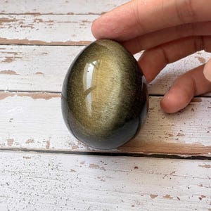Può includere: Una gemma lucida a forma di uovo con una lucentezza dorata e una base scura. La pietra ha una superficie liscia e riflettente e il colore dorato sembra turbinare al suo interno. La gemma è tenuta da una mano su uno sfondo di legno bianco e marrone.