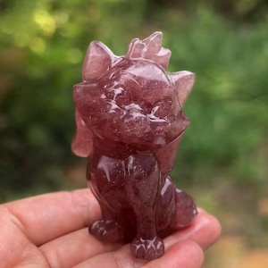 May include: A carved, reddish-purple crystal figurine of a cat. The cat is sitting upright with a bow on its head and a spiky design on the top of its head. The figurine is held in a person's hand against a blurred green background.